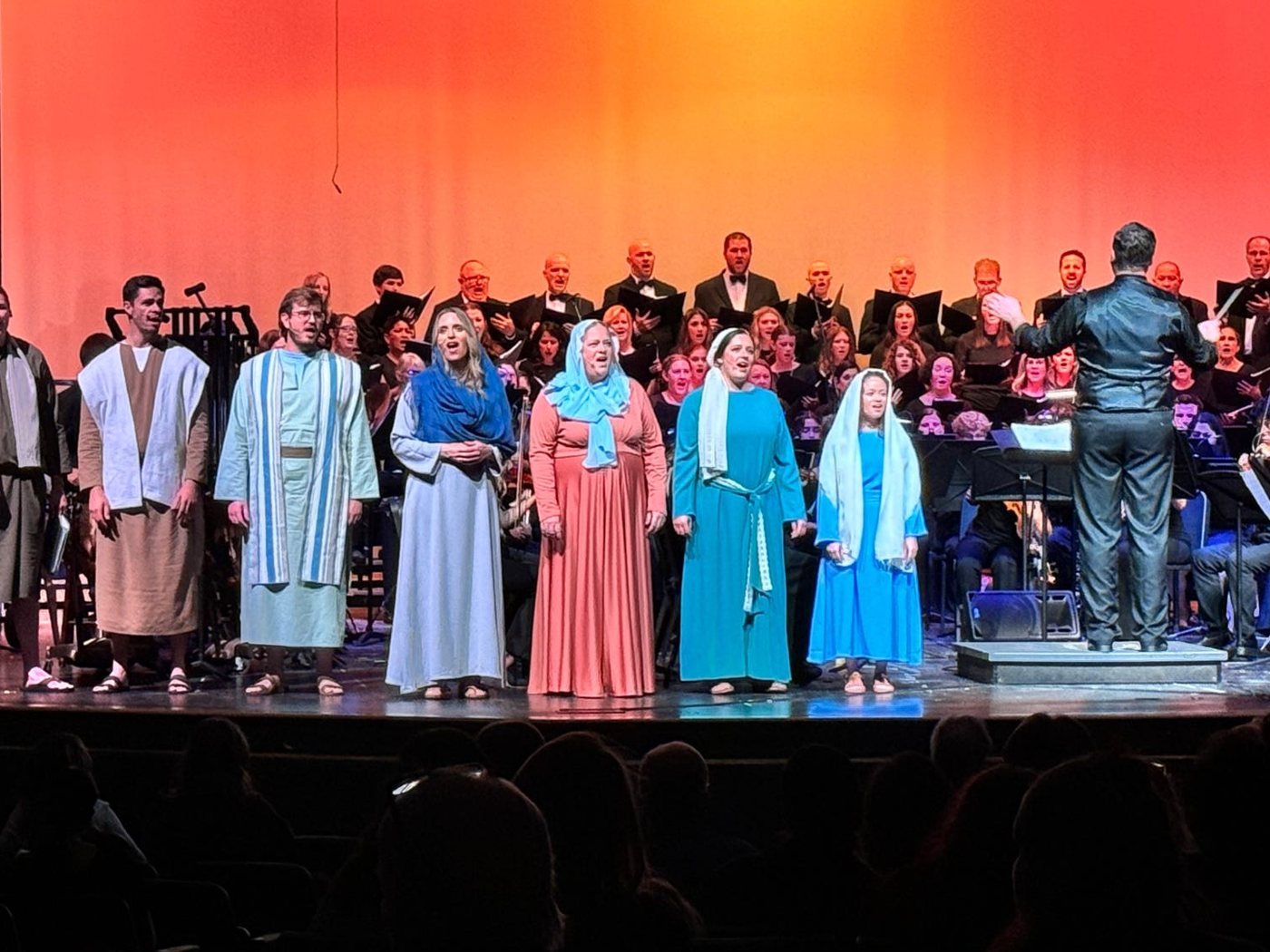 Soloists in costume standing in front of the choir and orchestra on stage during a Lamb of God performance