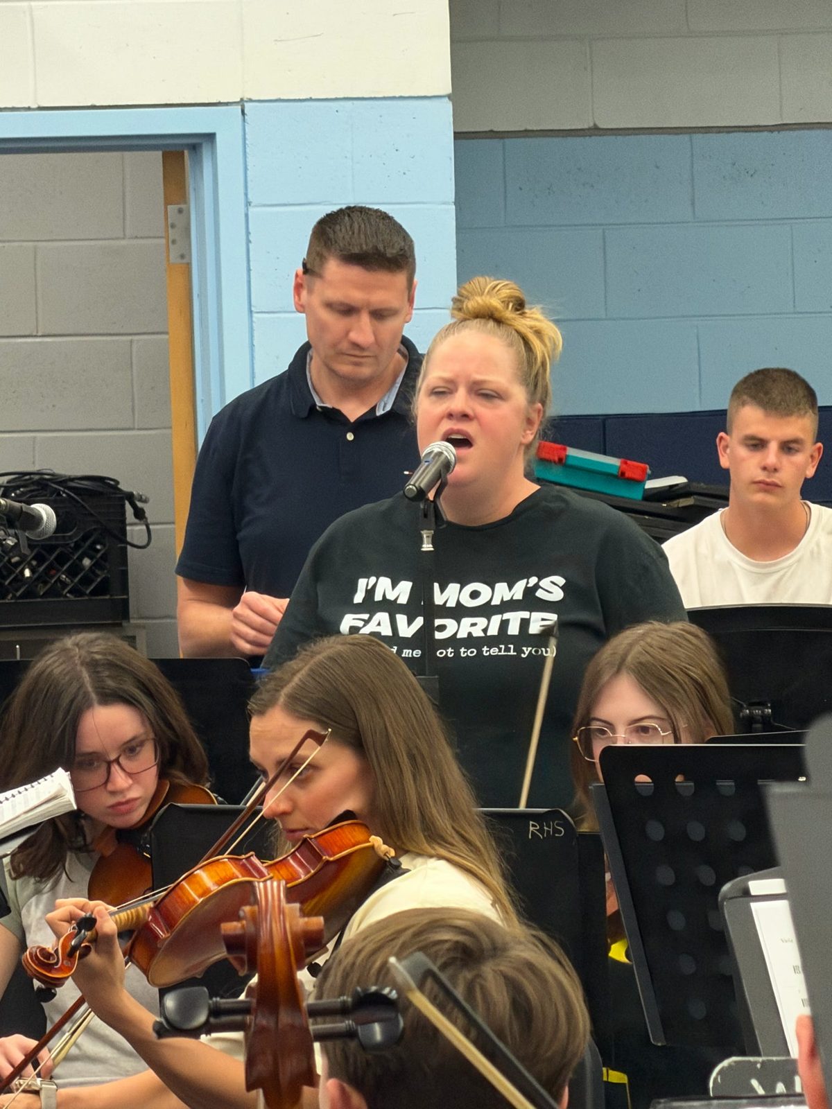 A singer at the microphone with young violinists in rehearsal